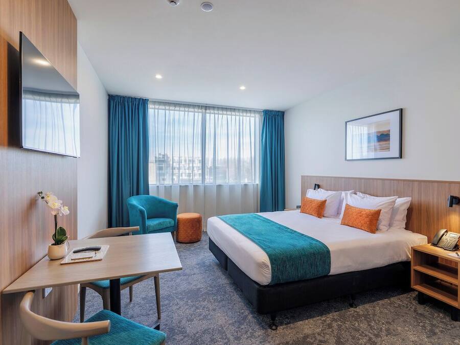 A hotel room with a bed, a desk, a chair, a small table, a nightstand, a phone, a framed picture, and a window with curtains.
