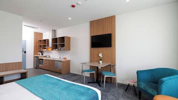 A modern hotel room with a bed, a desk, a chair, a sofa, a TV, and a kitchenette.