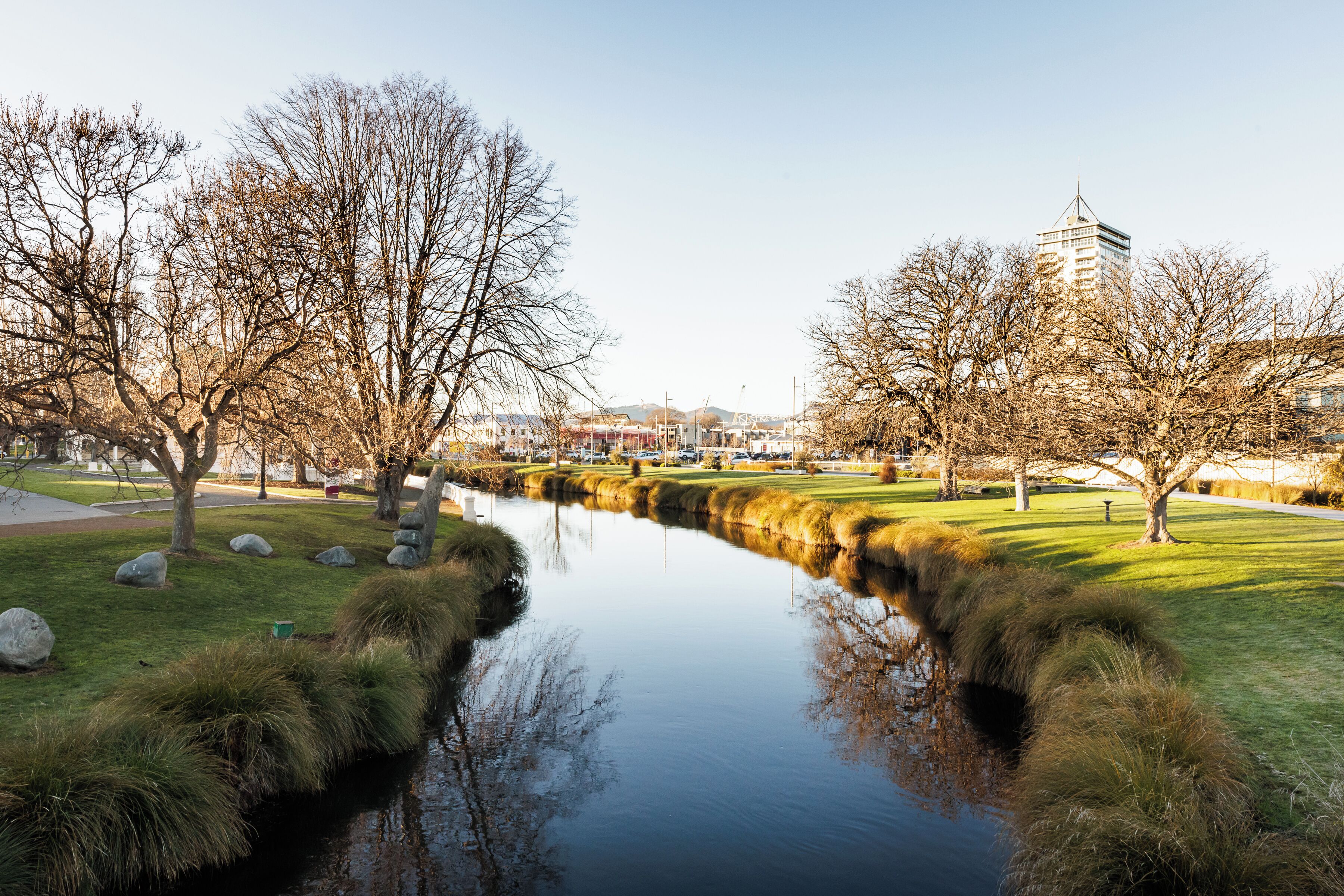 A serene park with a calm water feature, trees, and a distant building.