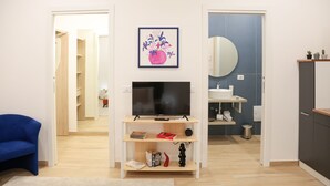 Una sala moderna con un televisor sobre un mueble de madera, un sillón azul, una obra de arte enmarcada y vistas al baño contiguo.