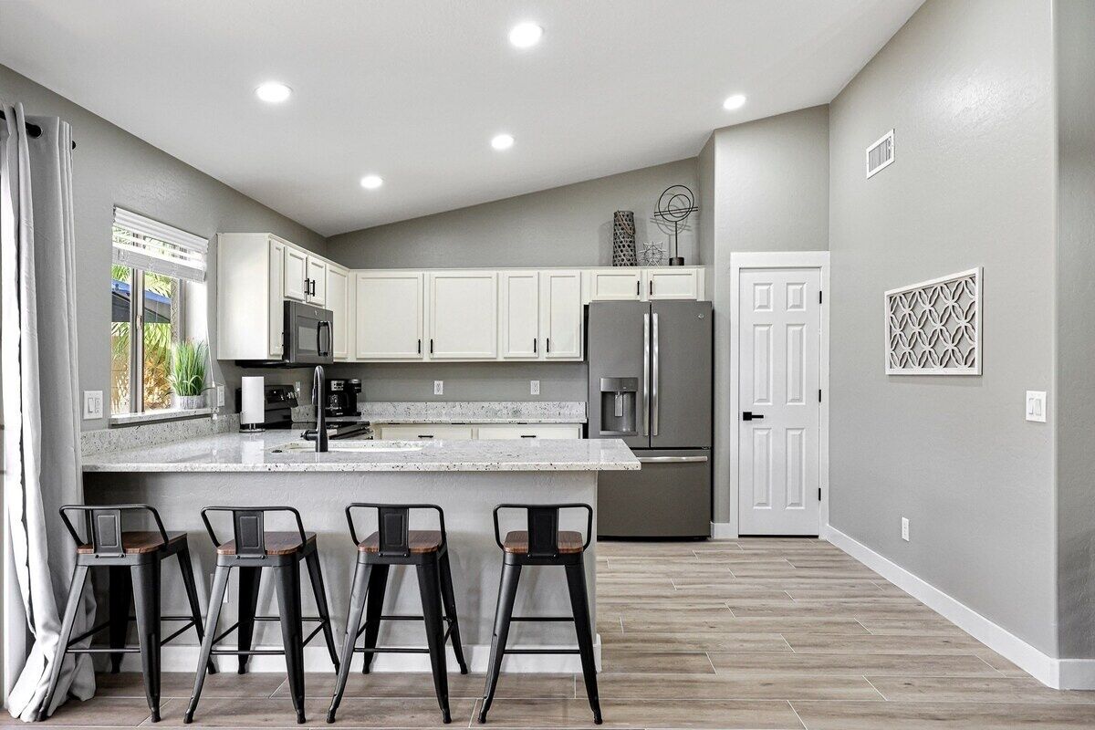 A modern kitchen with white cabinets, a large island, and stainless steel appliances. There are three bar stools and a door leading to another room.