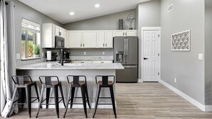 Cocina moderna con armarios blancos, una isla grande y electrodomésticos de acero inoxidable. Hay tres taburetes de bar y una puerta que da a otra habitación.