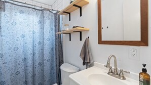 A bathroom with a shower curtain, toilet, sink, and a mirror. There are wooden shelves above the toilet and a towel hanging on the wall. - Charming Tiny Home Retreat!  (Austin)