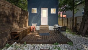 A small, dark blue building with a white door, a wooden deck, and a table with chairs. - Charming Tiny Home Retreat!  (Austin)