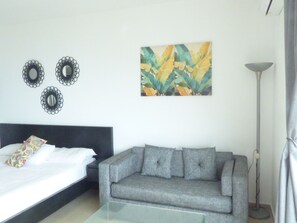 Ein Schlafzimmer mit einem Bett, einem grauen Sofa, einem Glastisch, einer Stehlampe und einem Bild an der Wand.