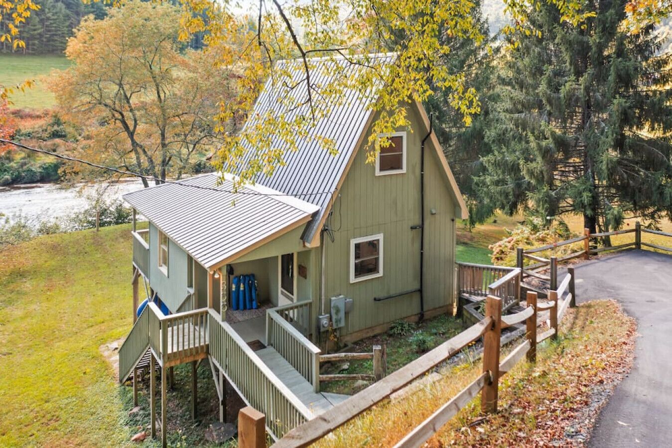 Een chalet met twee verdiepingen, een veranda en een trap, omringd door bomen en een rivier.