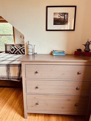 A bedroom with a bed, a dresser with drawers, a framed picture on the wall, and a window with a view of trees.