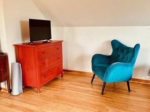 Une pièce avec un plancher en bois, une commode rouge sur laquelle est posée une télévision à écran plat, et un fauteuil turquoise.