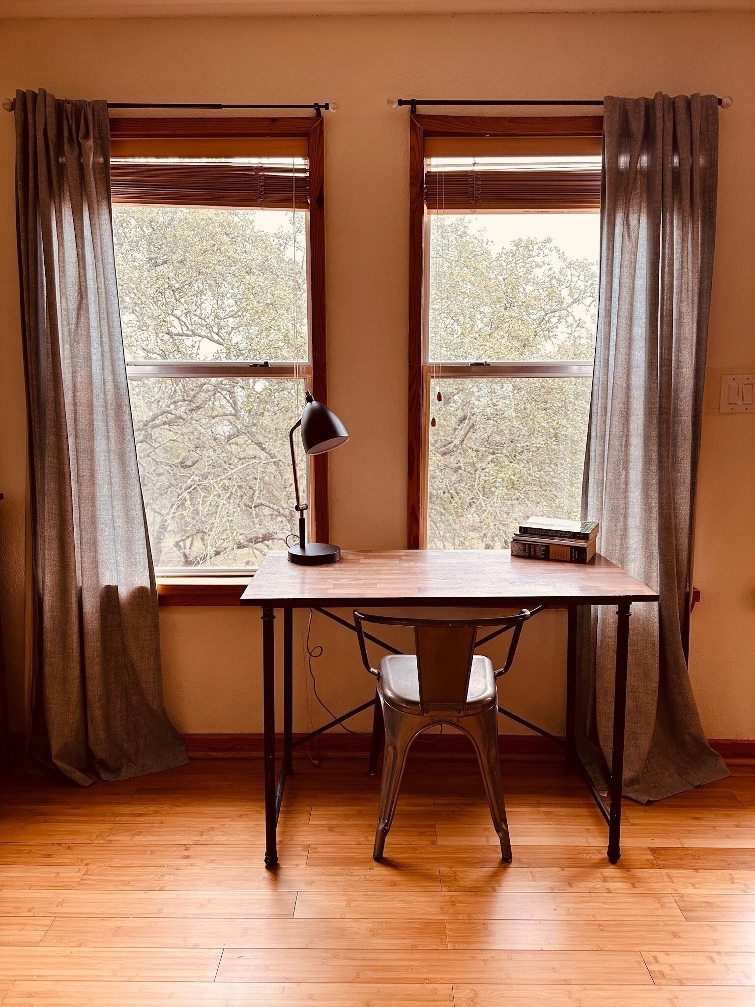 A room with a wooden desk, two windows with curtains, and a view of trees outside.