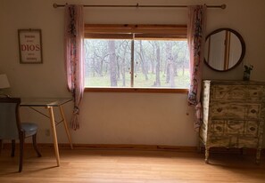 Una habitación con un ventanal grande, un escritorio de madera, una silla, un aparador y un espejo en la pared.