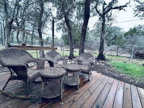 Una terraza de madera con muebles de mimbre, incluyendo sillas y una mesa, rodeada de árboles.