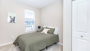 A bedroom with a bed, a window with blinds, a wall-mounted picture, and a white door.