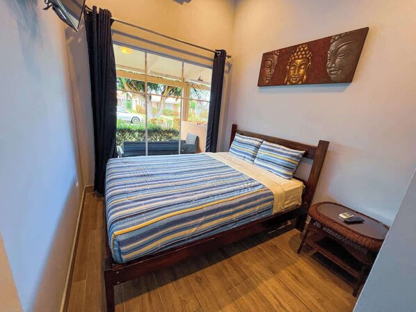 A bedroom with a bed, a window with curtains, a small table, and a wall-mounted artwork.