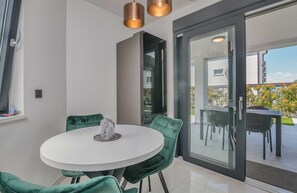 Et moderne spiseområde med et rundt bord og grønt polstrede stole, en skydedør, der fører til en udendørs terrasse, og hængende lamper.