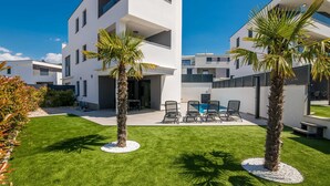 Een modern woongebouw met twee verdiepingen, een plat dak, een balkon en een klein terras met stoelen en een tafel.