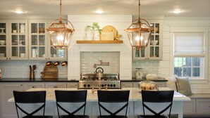 Una cocina con muebles blancos, una isla central con taburetes de bar, una estufa y lámparas colgantes.