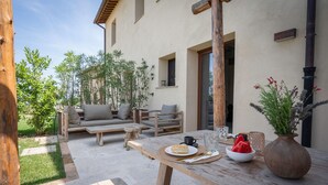 An outdoor patio with a wooden table set for dining, a sofa, and a chair. There are plants and a small garden area visible. - Beautiful apartment with pool, WIFI, A/C, TV, veranda and panoramic view, close to San Gimignano (Montaione)