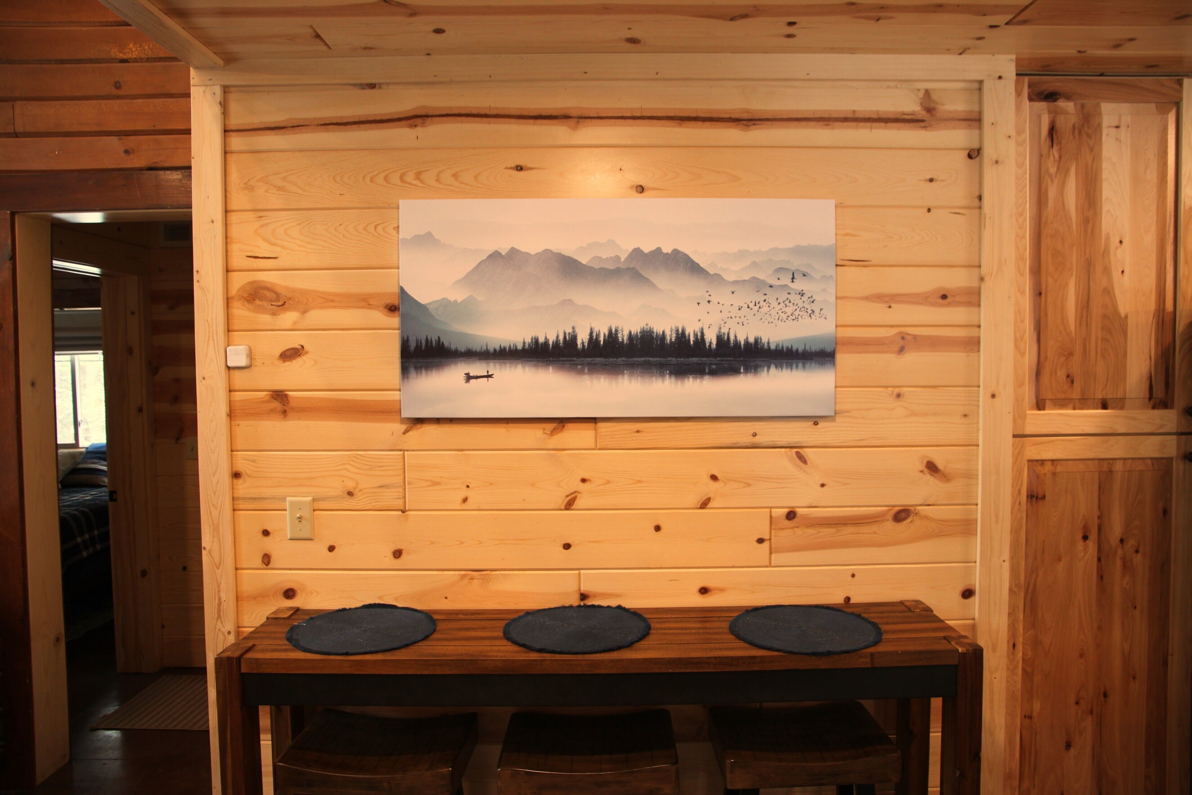 A wooden cabin interior with a dining table, benches, and a painting of a mountain landscape.
