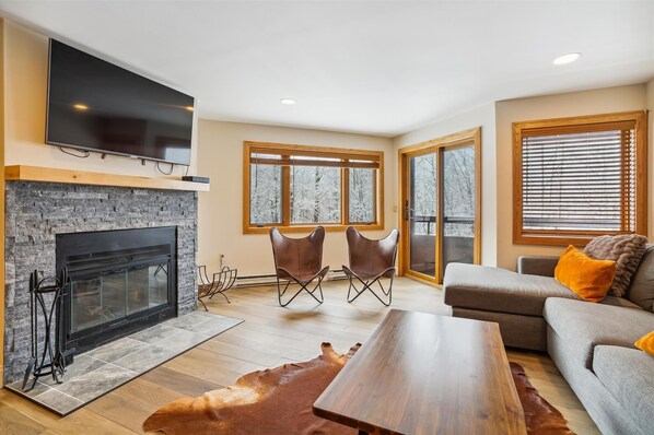 A modern living room with a fireplace, a flat-screen TV mounted above it, a sofa with orange cushions, and a wooden coffee table. - 2 bedroom condo with mountain views, pool, hot tub, and private sauna. (Killington)