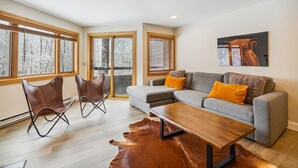 A modern living room with a grey sofa, two brown leather chairs, a wooden coffee table, and a large window with a view of snow-covered trees. - Two-Bedroom Condo with Stunning Views and Prime Location A4 (Killington)