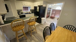 Una cocina compacta con armarios blancos, un refrigerador negro, una mesa de madera con cuatro sillas y un cubo de basura a la vista.