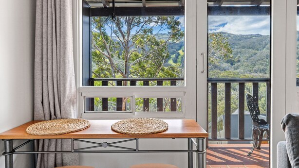 A balcony with a view of trees and hills, a table with woven placemats, and a chair.