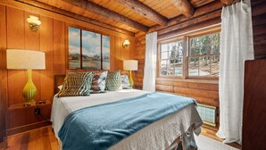 Un dormitorio de cabaña de madera con una cama, dos lámparas de pared, una ventana con cortinas y vistas a árboles.