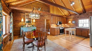 Interior de una cabaña de madera con una mesa de comedor, sillas, una cocina con electrodomésticos y un amplio ventanal.