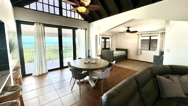 A spacious living area with a balcony overlooking a scenic view, a dining table with chairs, and a cozy seating area. - Endless ocean views and pool, by El Yunque. (Río Grande)