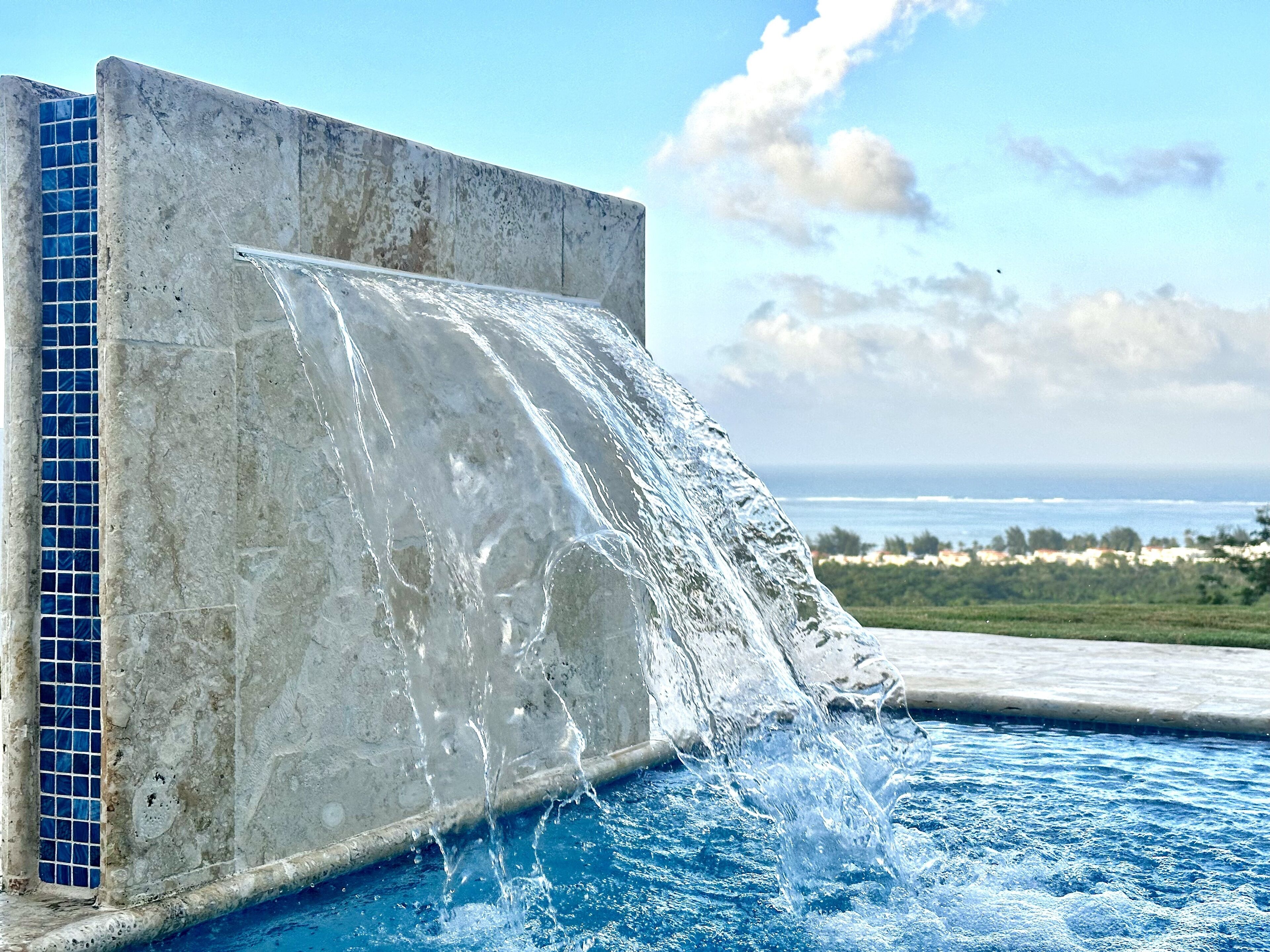 A pool with a waterfall feature, clear blue water, and a view of the ocean and distant buildings.