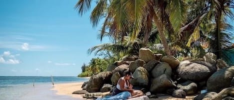 A beach with clear water, a person lying on a mat, and palm trees.