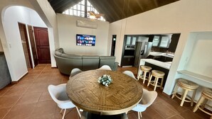 A modern kitchen with a dining area, featuring a round wooden table with chairs, a sofa, and a TV mounted on the wall. - Endless ocean views and pool, by El Yunque. (Río Grande)