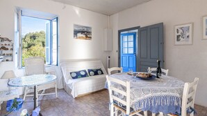 Een kamer met een bed, een eettafel met stoelen, een raam met uitzicht op bomen, en een blauwe deur.