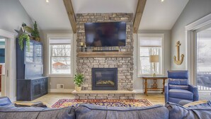 A living room with a stone fireplace, a flat-screen TV mounted above it, a blue leather sofa, and a large window with a view of a house. - Blue Skies - Lakefront Living at it's Finest! Luxury Amenities! (Honeoye)