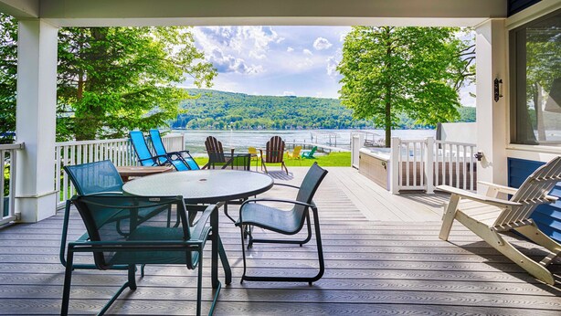 A covered outdoor deck with a table and chairs, overlooking a lake and forested hills. - Blue Skies - Lakefront Living at it's Finest! Luxury Amenities! (Honeoye)