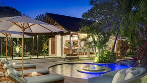 Zona de piscina con sillones de descanso y sombrillas, un comedor y un edificio bien iluminado con piscina.
