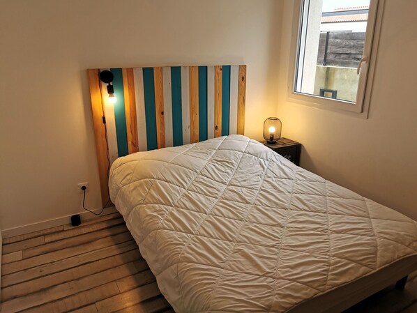A bedroom with a wooden headboard, a white quilted bedspread, a bedside table with a lamp, and a window with a view of the outside.