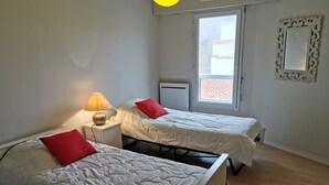 A room with two beds, each with a red pillow, a bedside table with a lamp, and a window with a view of buildings. - The Dunes of Blossac 100m from the beach (Les Sables-d´Olonne)