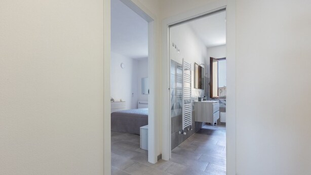 Un couloir mène à une chambre à coucher qui contient un lit, une petite table de chevet et une commode. On aperçoit une salle de bain avec des toilettes, un lavabo et un miroir. Le sol est carrelé et les murs sont peints en blanc.