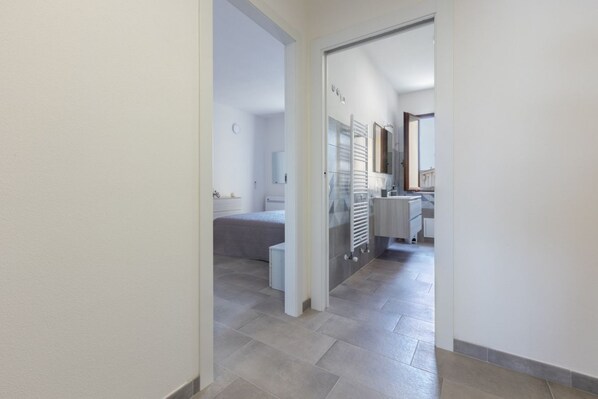 A hallway leading to a bedroom with a bed, a small bedside table, and a nightstand. There is a bathroom visible with a toilet, sink, and mirror. The floor is tiled, and the walls are painted white.