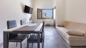 Une pièce moderne comprenant une table à manger, des chaises, un canapé, un téléviseur fixé au mur et une fenêtre donnant sur des immeubles.