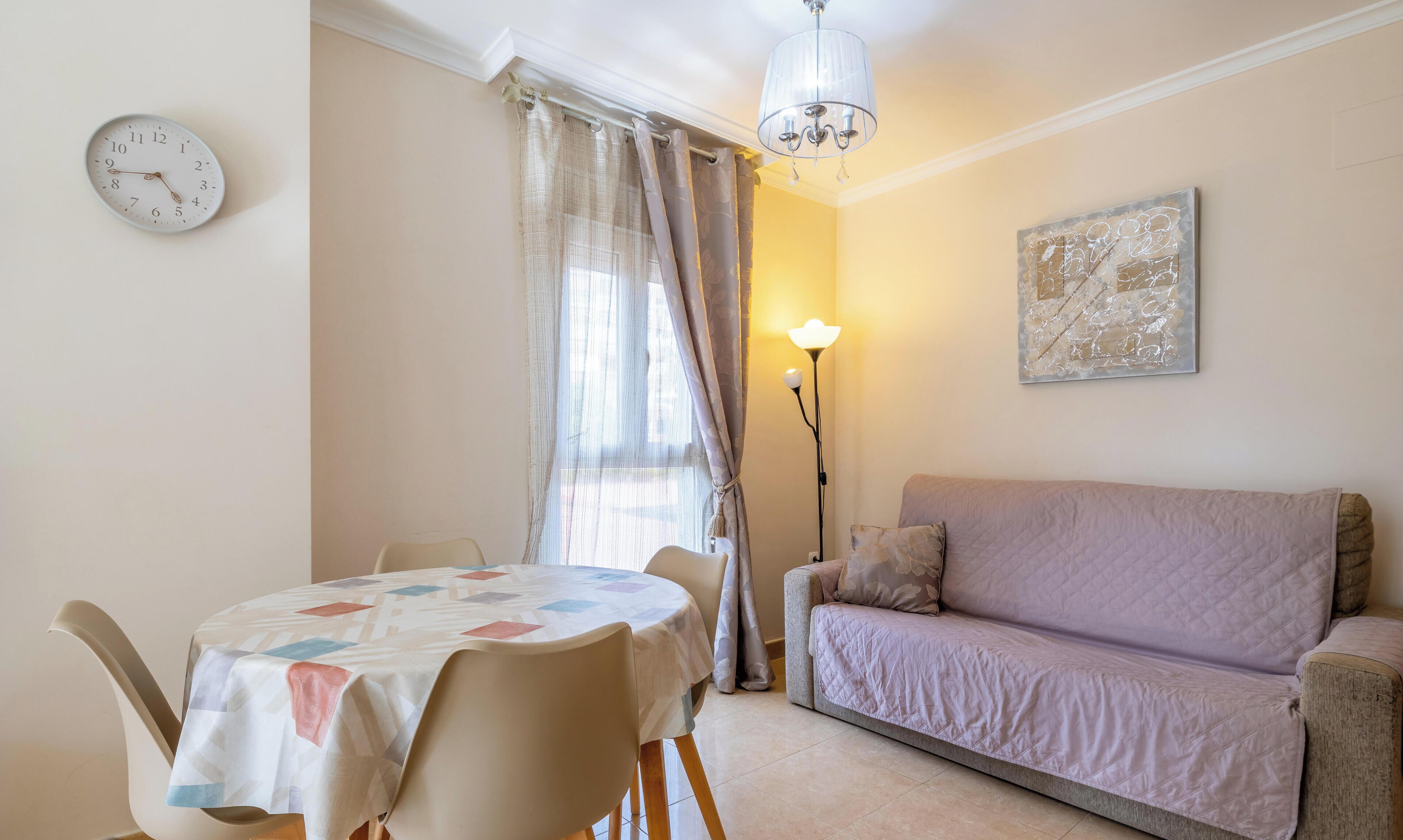 Un salon avec un canapé, une table entourée de chaises, une horloge, une lampe et une fenêtre avec des rideaux.