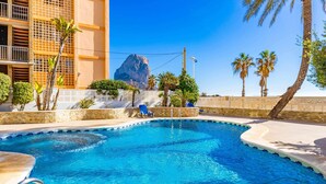 A swimming pool with clear blue water, surrounded by palm trees and a building with a rock formation in the background. - Inftour Porto Azul 1 Apartamento (Calp)