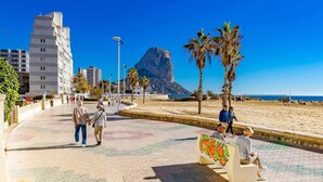 A tiled walkway with palm trees and benches, a mountain in the background, and a clear blue sky. - Inftour Porto Azul 1 Apartamento (Calp)