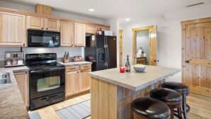 A modern kitchen with wooden cabinets, a black stove, a refrigerator, and a bar area with stools.