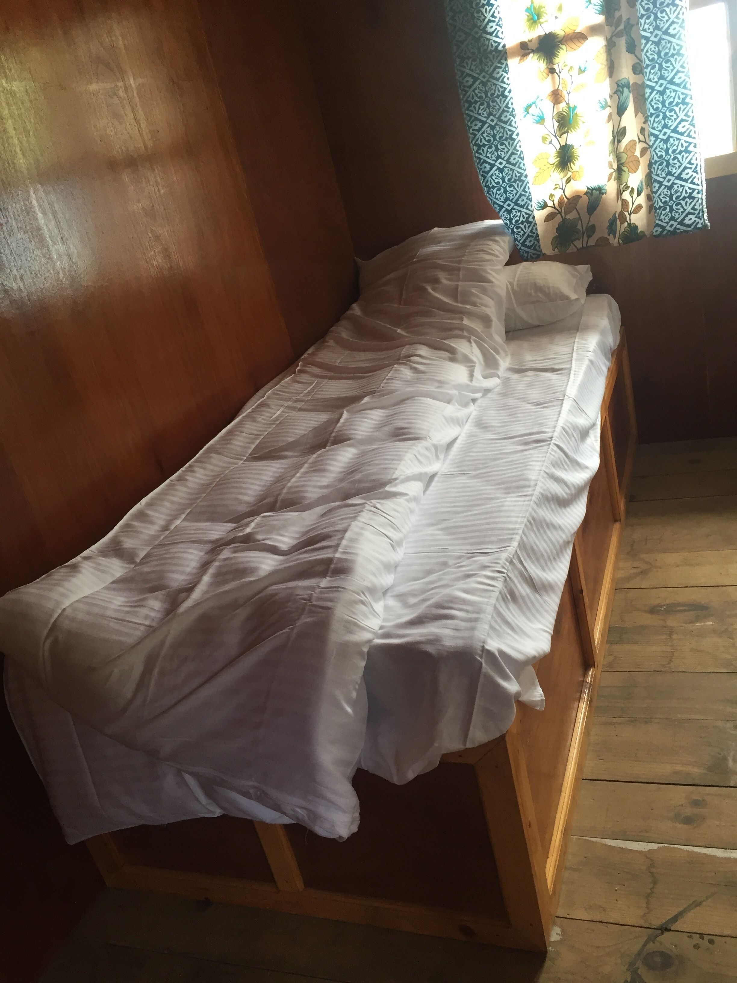 Una cama de madera con ropa blanca en una habitación con suelo de madera y una ventana con cortinas estampadas.