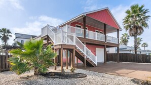 A two-story house with a red exterior, a balcony with white railings, and a wooden deck. - Corral Cottage- Private Pool Oasis, Games, Pets, Golf Cart zoned & Walk to Beach (Port Aransas)