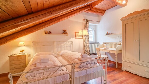 A bedroom with a sloped ceiling, a bed with white linens, a nightstand with a lamp, a dresser, and a window with curtains. - La Torre In The Center Of Town, Sauze d'Oulx, Italy (Sauze d'Oulx)