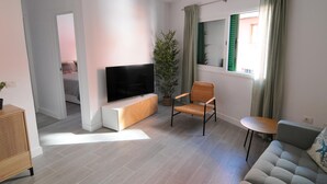 Una sala de estar moderna con un televisor de pantalla plana sobre un mueble blanco, una silla de mimbre, un sofá gris y una mesita.