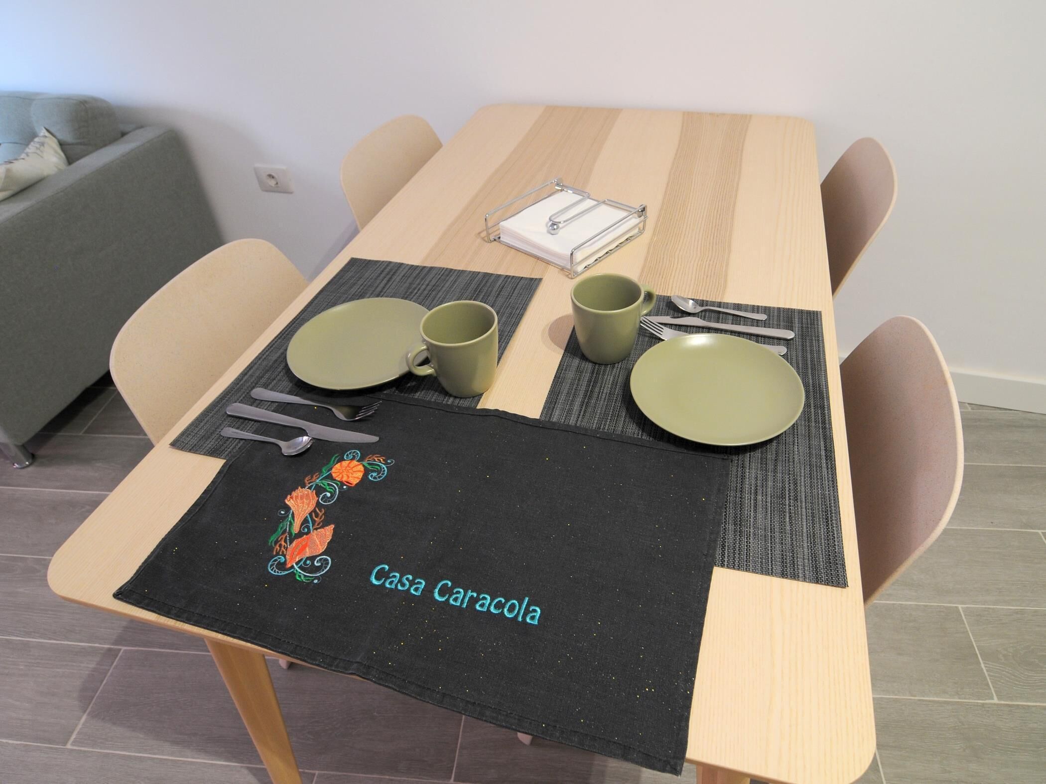 A dining table set with plates, cups, and cutlery, a placemat with 'Casa Caracola' text, and a chair.